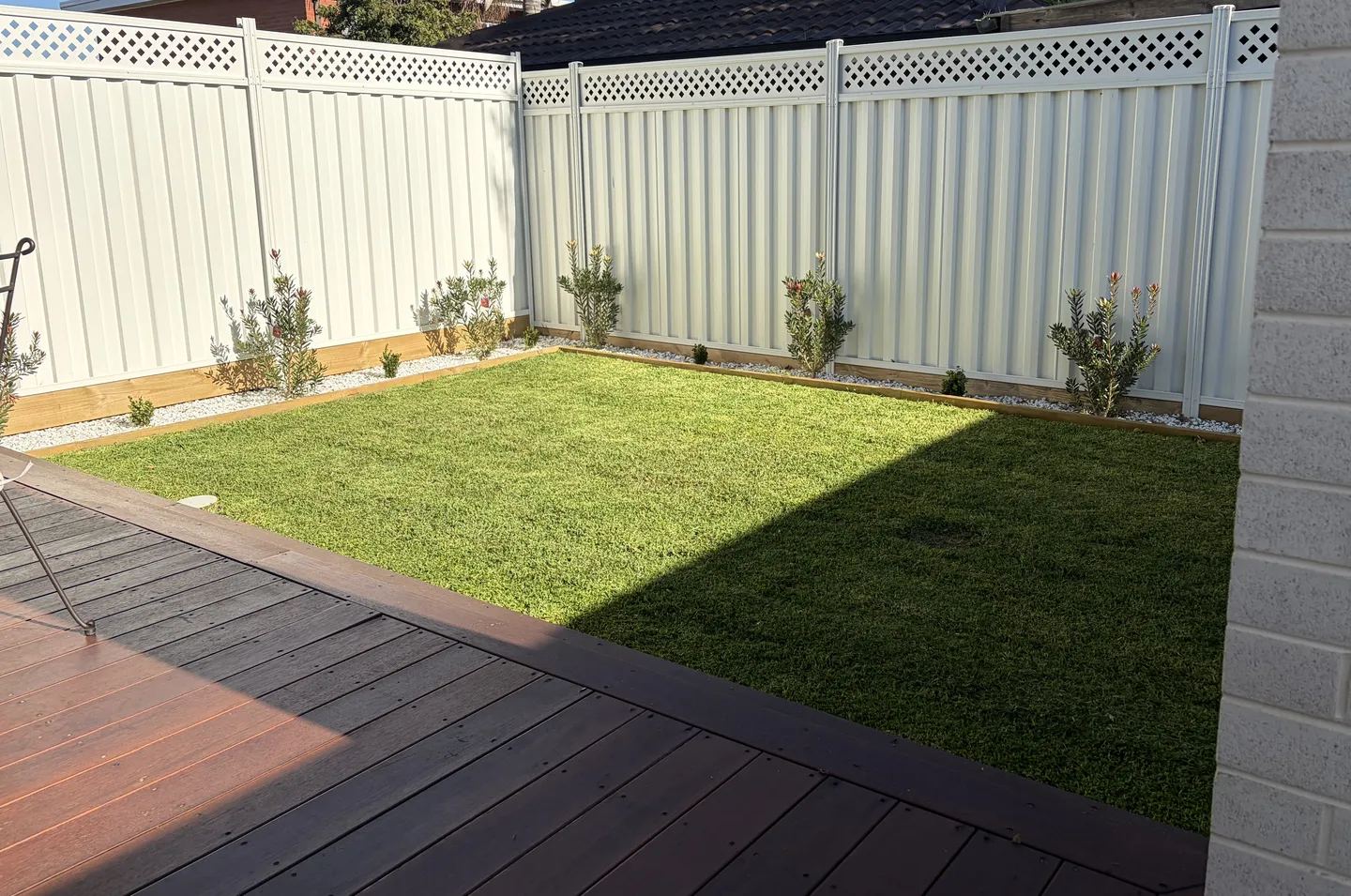 A green backyard lawn mowed neat and short, bordered by a timber deck and small garden beds along a white fence